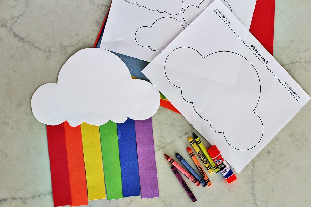 Cloud cutout with paper strips to make a rainbow next to printable pages of clouds and a small pile of crayons by a gluestick.