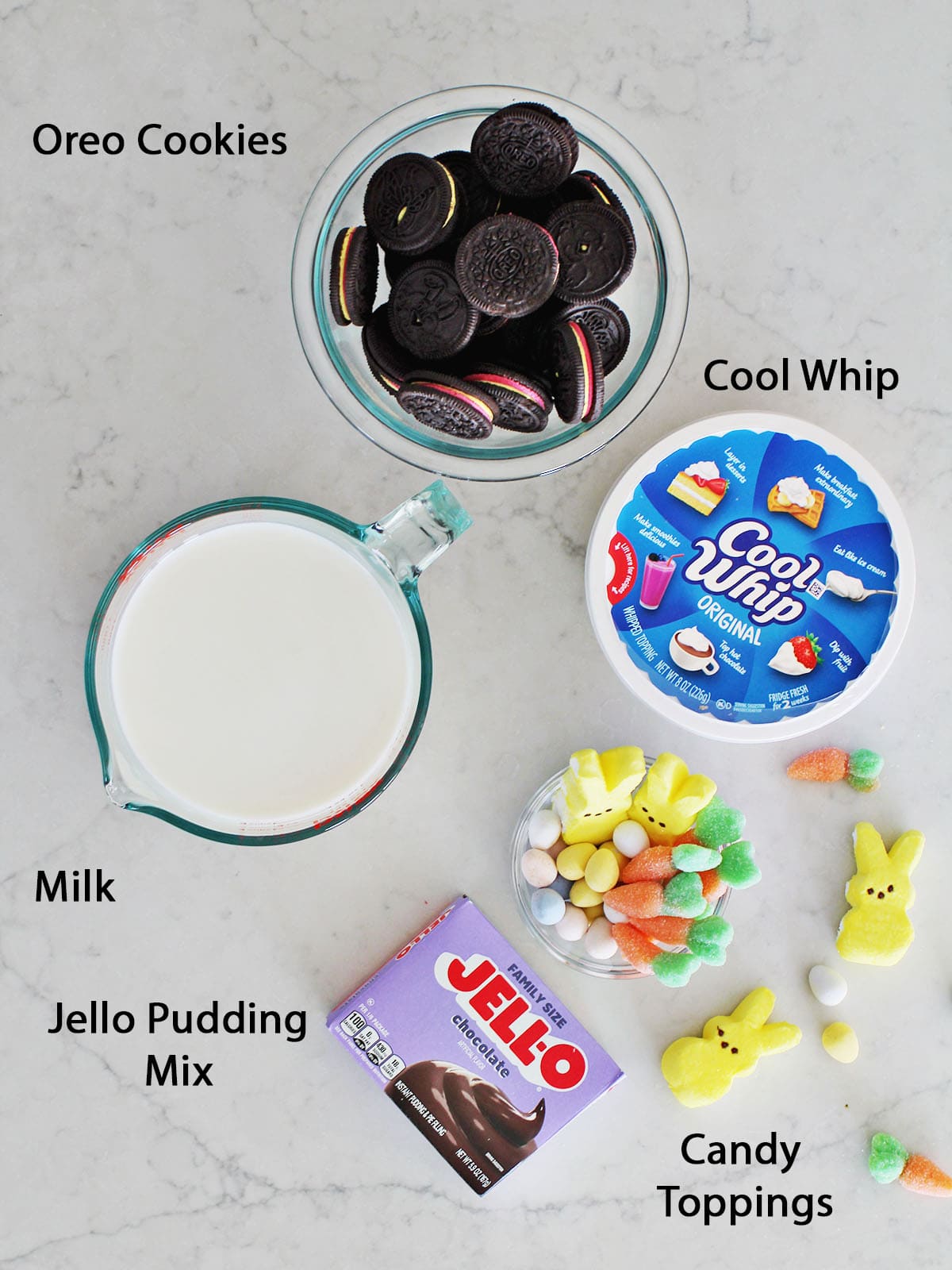 Bowl of oreos, container of cool whip, measuring bowl of milk, a box of chocolate pudding mix and Easter candy in a bowl on a counter.