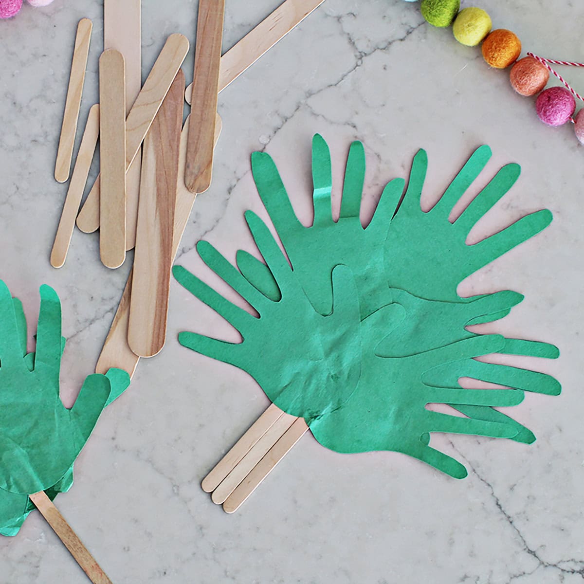Green cut out handprints glued to popsicle sticks to make a palm branch next to a pile of popsicle sticks in various sizes.
