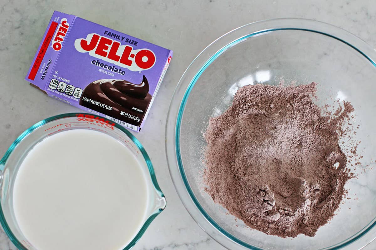Box of jello pudding mix, a measuring bowl of milk and pudding mix in a large clear bowl on a counter.