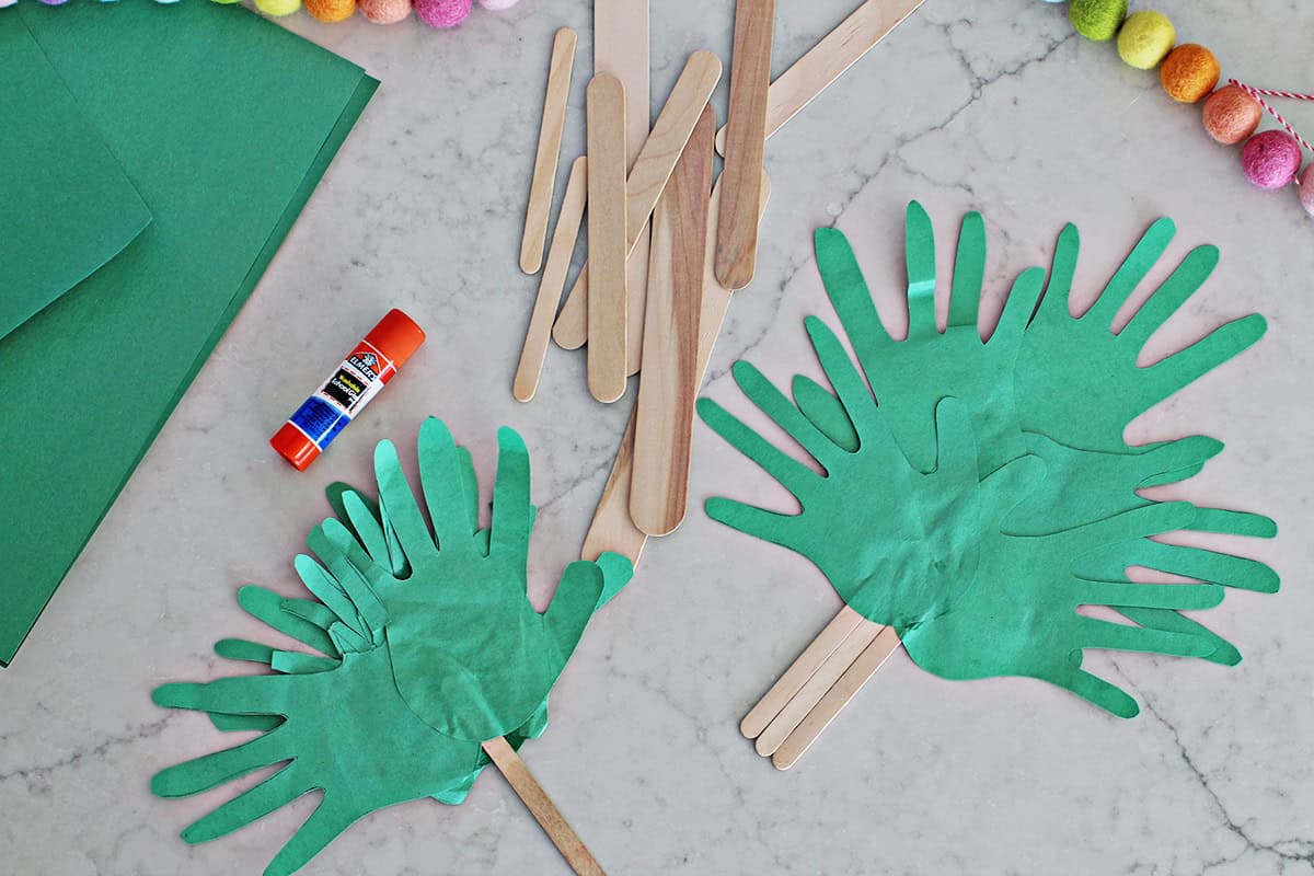 Green cut out handprints glued to popsicle sticks to make a palm branch next to a pile of popsicle sticks in various sizes and glue sticks.