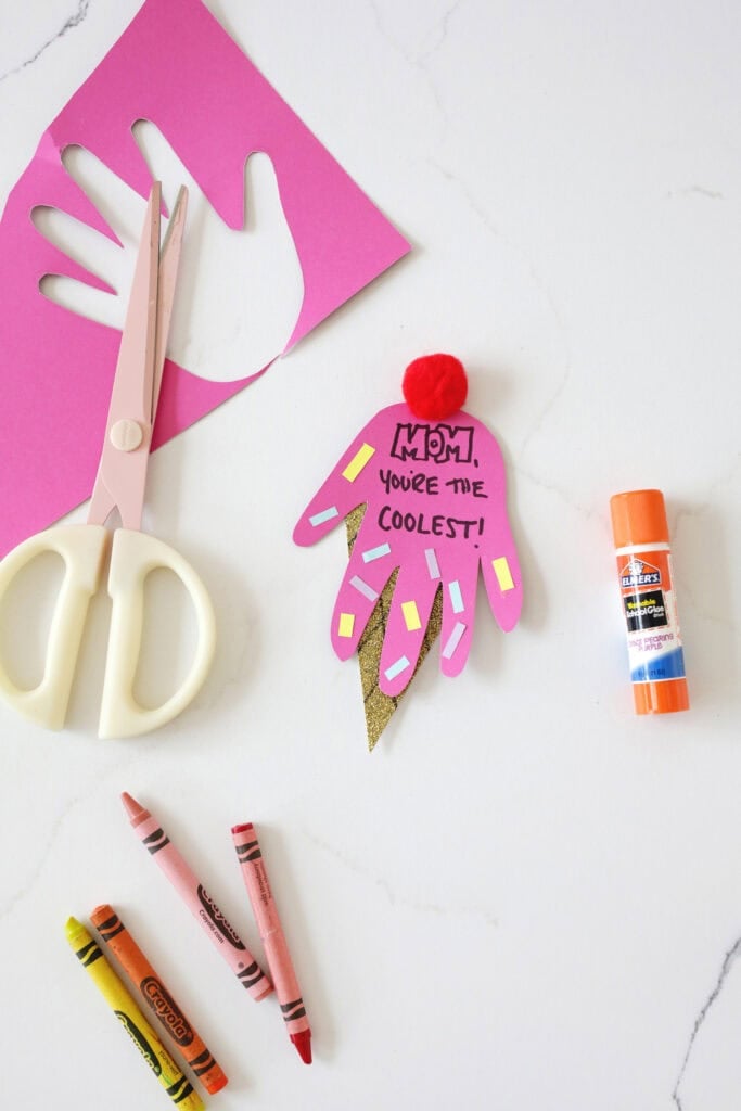 Pink construction paper next to a pair of scissors, a glue stick, crayons, and a handprint made to look like an ice cream cone.
