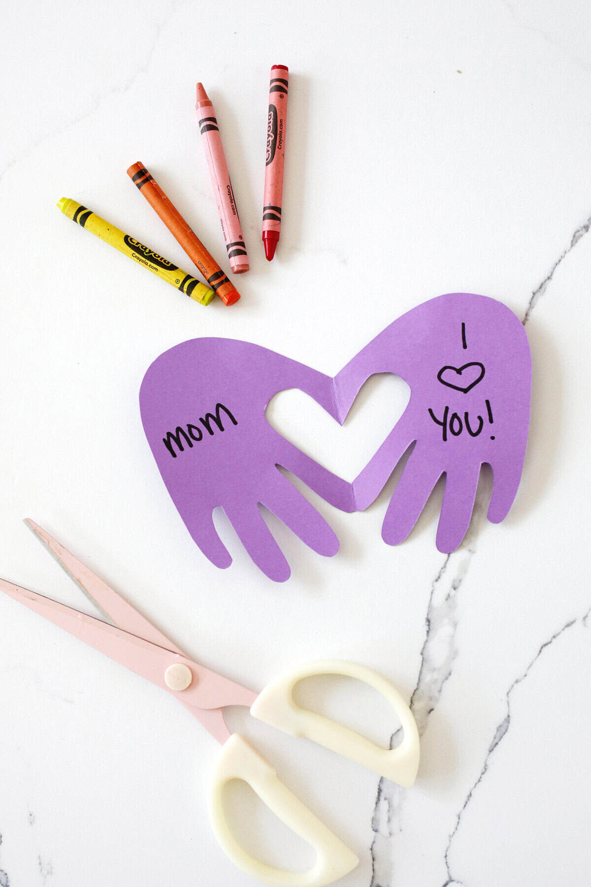 Handprint heart Mother's Day craft made from purple paper next to crayons and a pair of scissors.