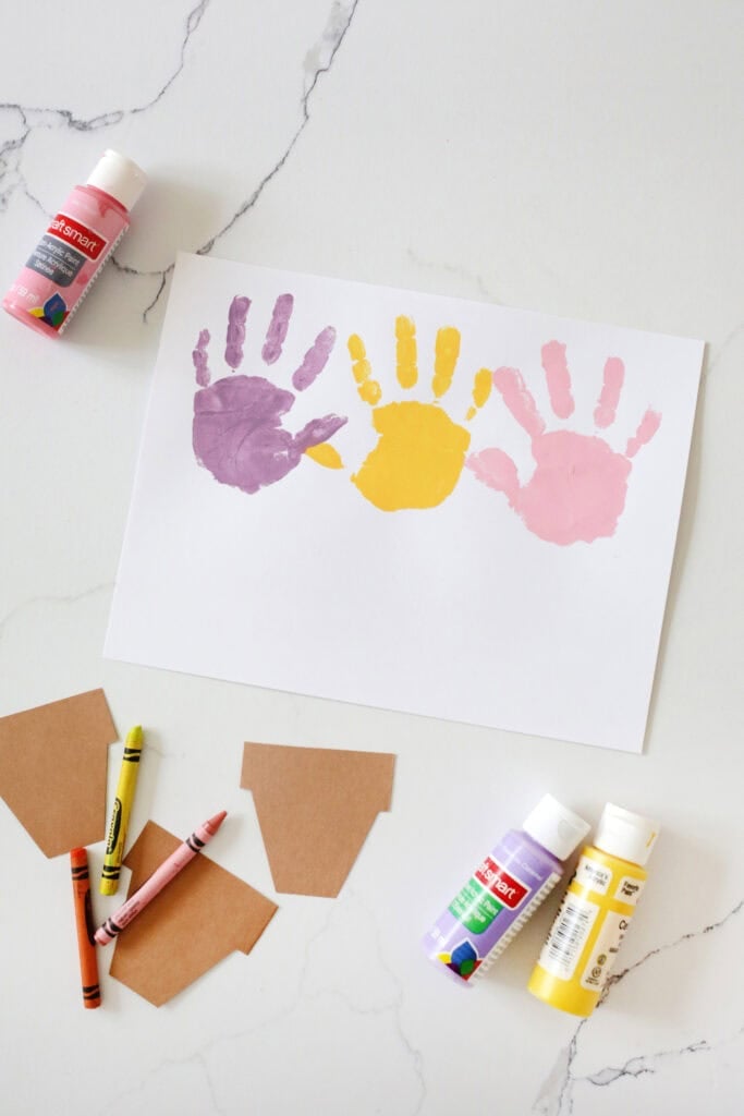 A purple, yellow and pink handprint on white paper next to cut out pots from brown paper.