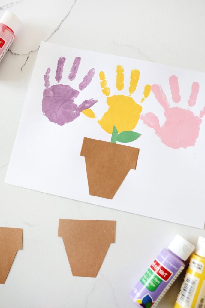 A purple, yellow and pink handprint on white paper next to cut out pots from brown paper with green stems to make the handprints look like flowers.