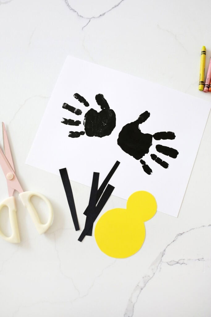 Two black handprints on a piece of paper next to cut out yellow shape and black strips of paper.
