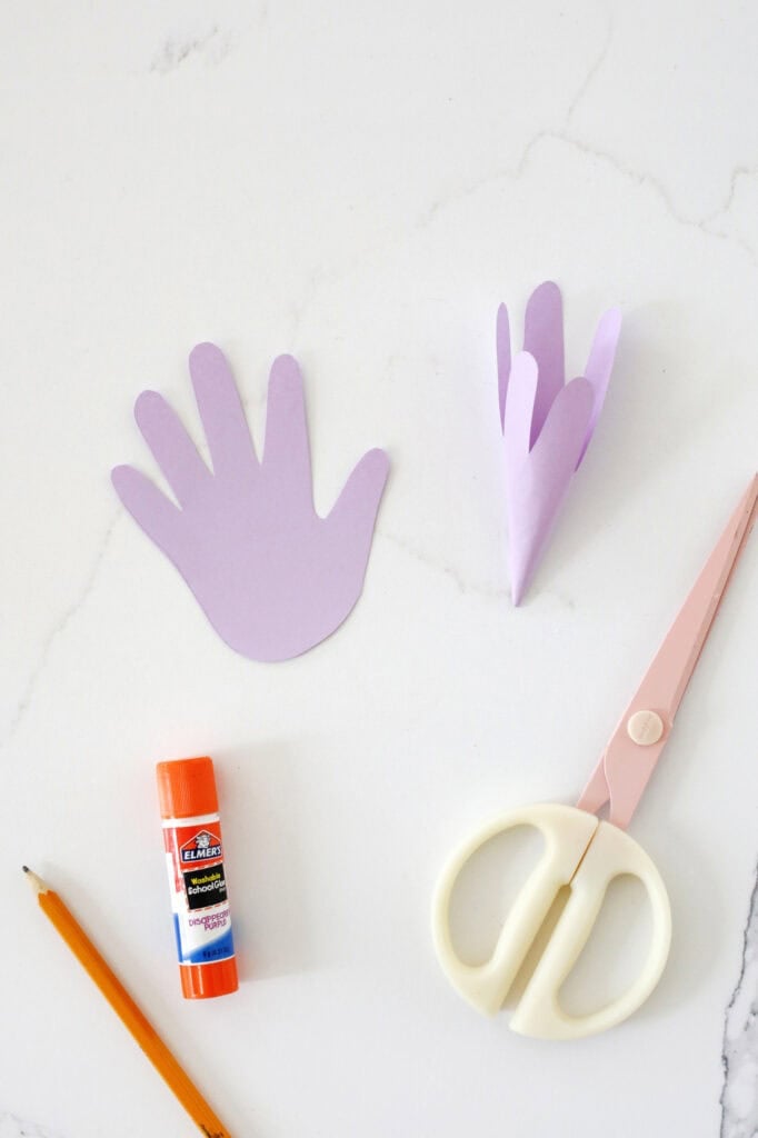 Purple paper cut into handprints to make a lily shape next to a gluestick, scissors and a pencil.