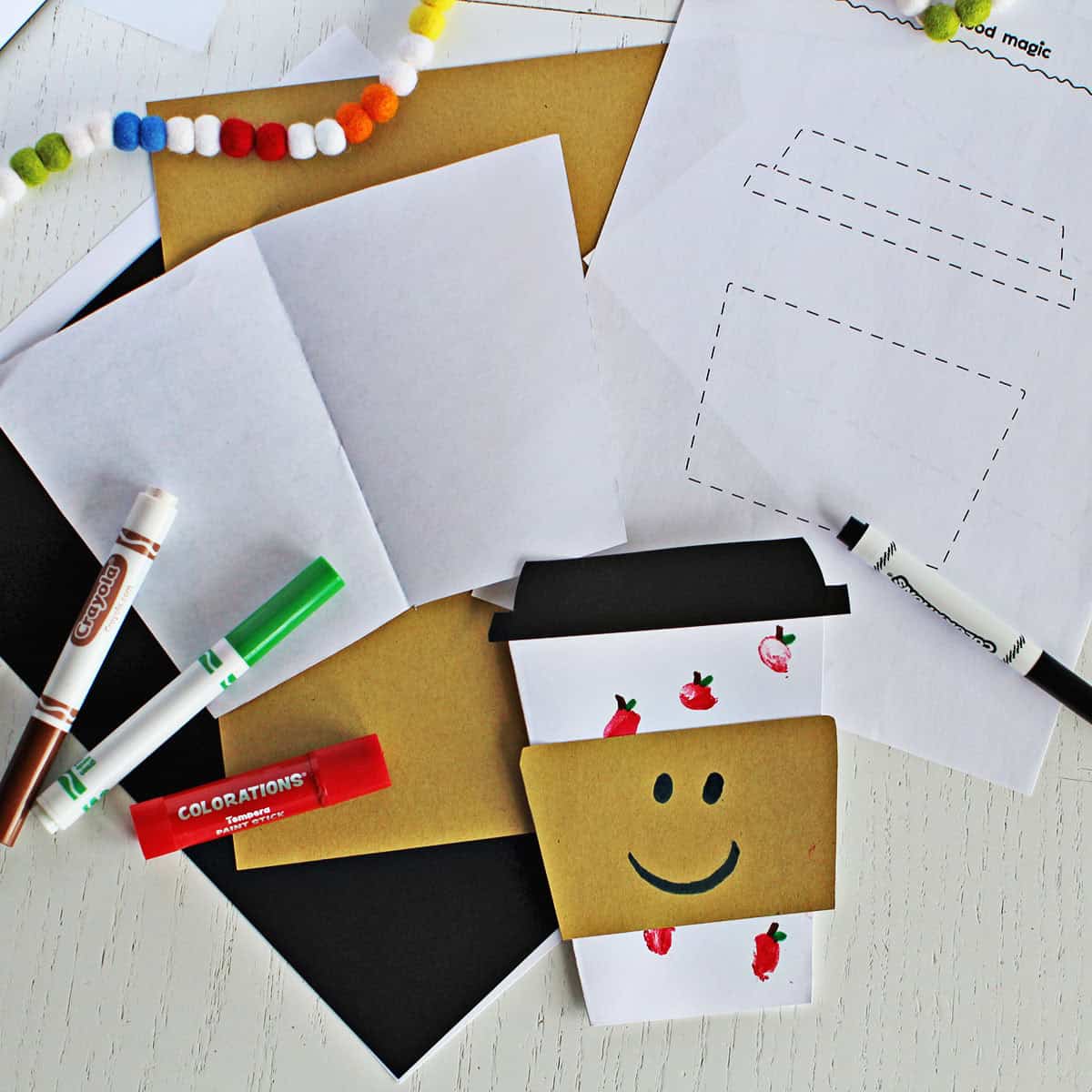 paper card shaped like a coffee cup with fingerprints made to look like apples next to the printed template and markers.