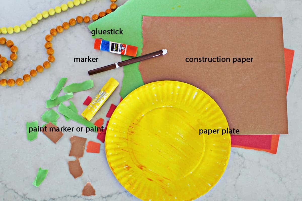 a paper plate that's been painted yellow, construction paper in brown, red, orange and gree, a brown marker and a yellow paint marker on a counter.
