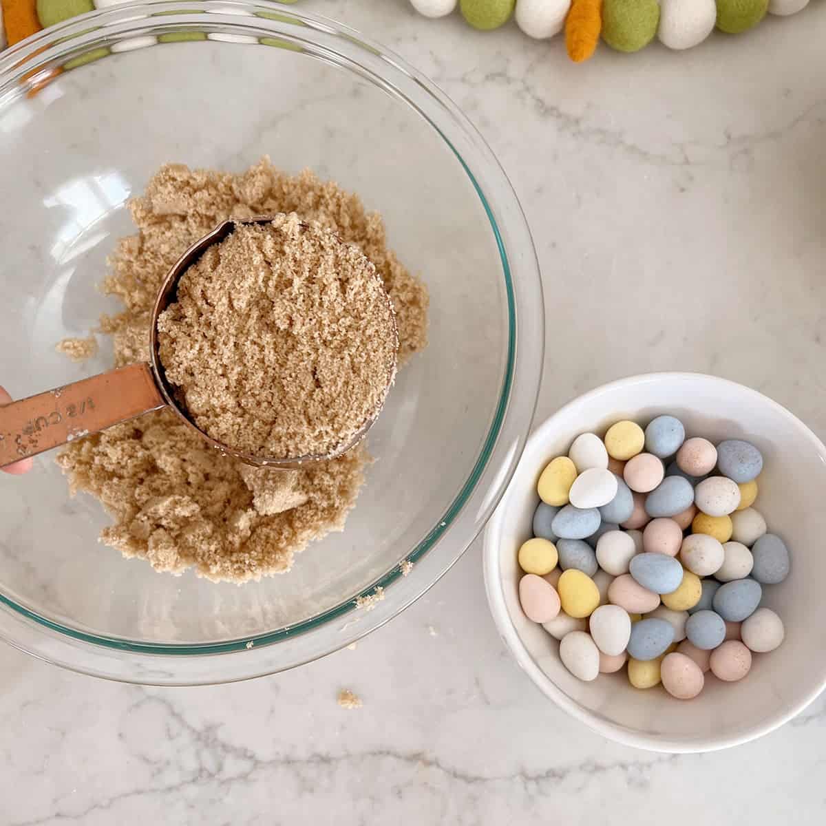 A measuring utensil filled with brown sugar being added to a bowl sitting next to a bowl filled with cadbury eggs.