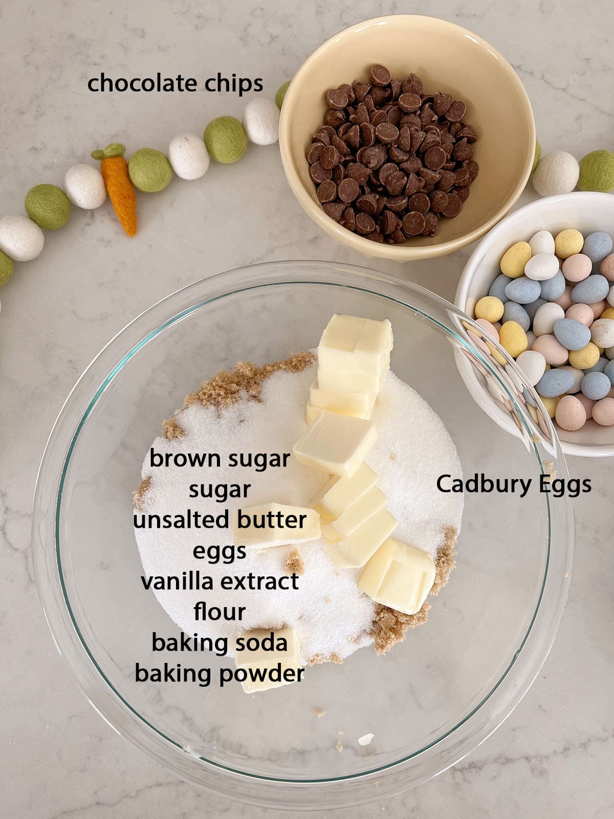 A bowl of chocolate chips, a bowl of cadbury eggs, and a mixing bowl with brown sugar, sugar and butter in it.