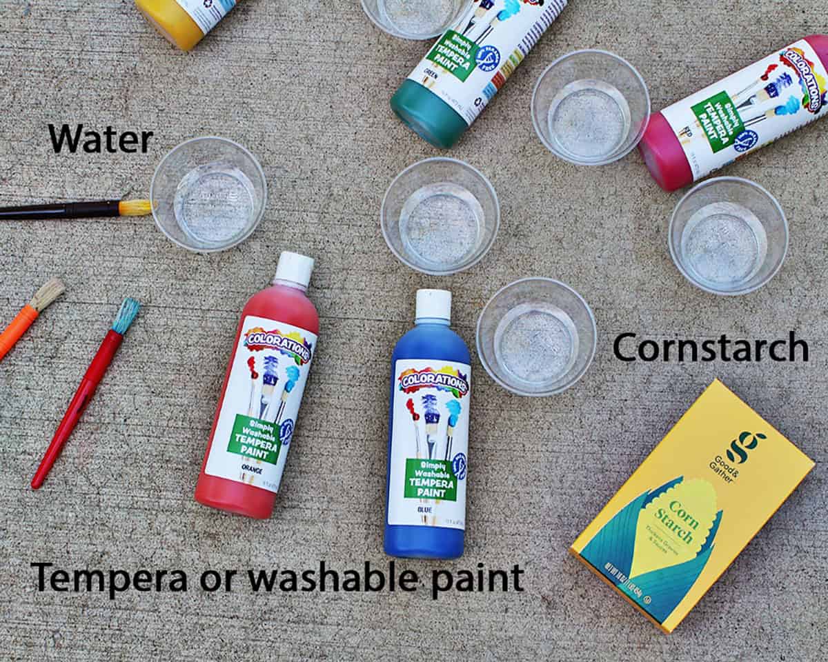 tempera paint in different colors on the ground next to paintbrushes, cups of water and a box of cornstarch.