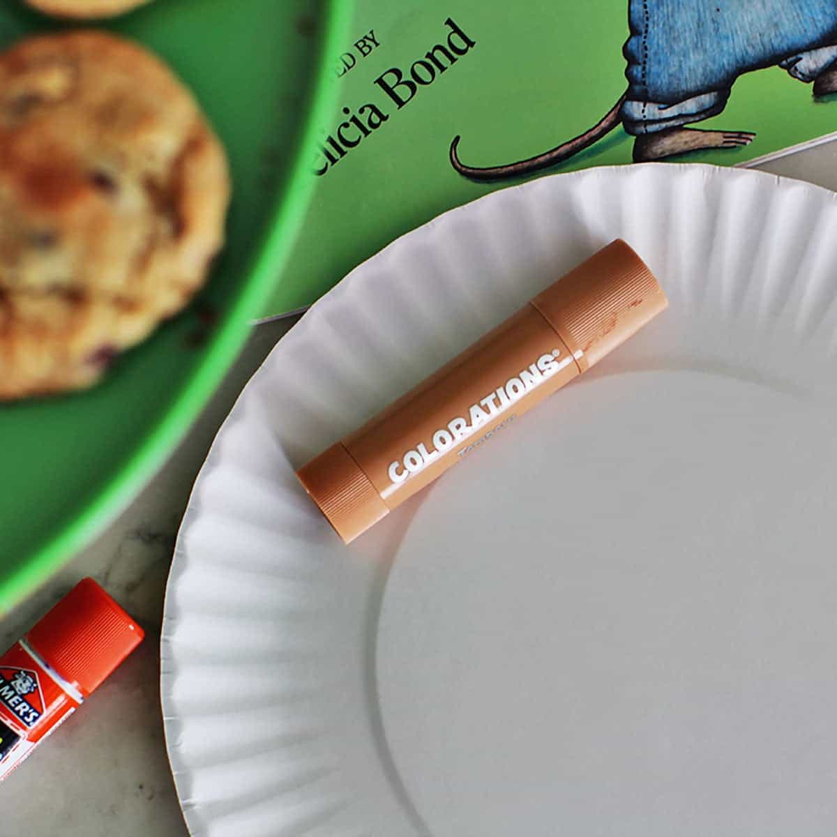 A plain paper plate with a paint marker sitting on it next to a glue stick, a book and a plate of cookies.