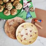 paper plates decorated to look like chocolate chip cookies next to a book, paper, gluestick, paint stick and a plate of cookies.