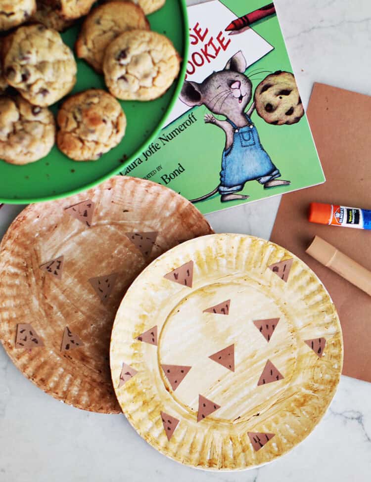 paper plates decorated to look like chocolate chip cookies next to a book, paper, gluestick, paint stick and a plate of cookies.
