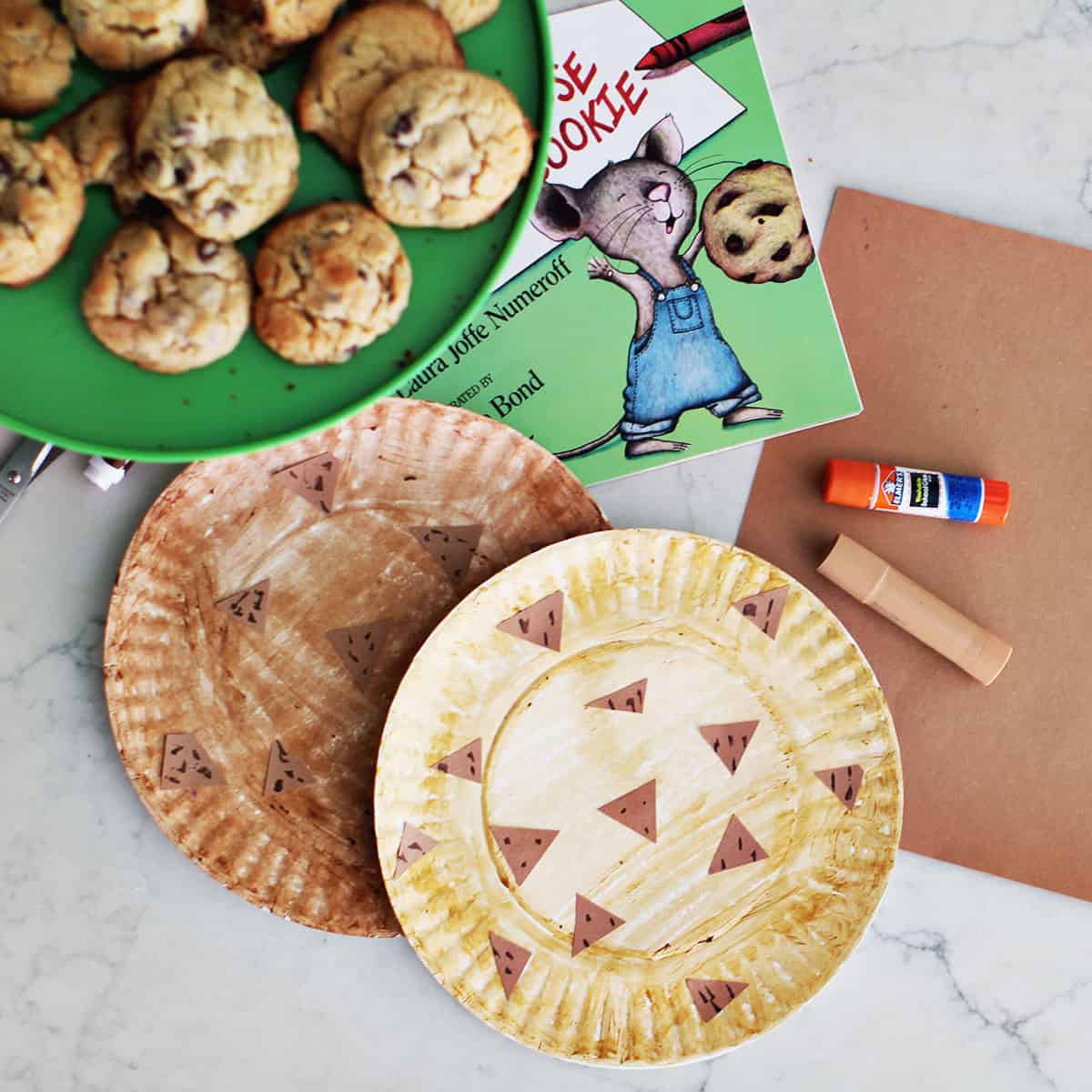 paper plates decorated to look like chocolate chip cookies next to a book, paper, gluestick, paint stick and a plate of cookies.