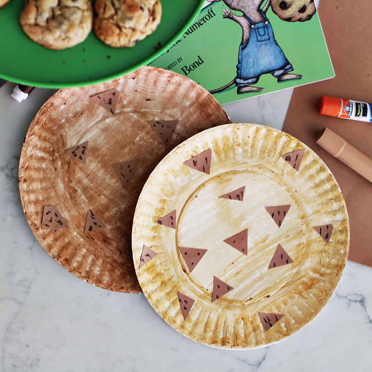 paper plates decorated to look like chocolate chip cookies next to a book, paper, gluestick, paint stick and a plate of cookies.