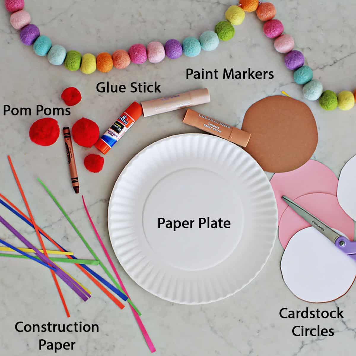 paper plate, strips of construction paper, cardstock circles, a glue stick, red pom poms and paint markers on a counter.