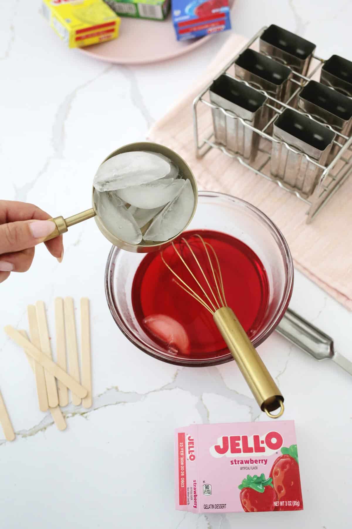 cup of ice being added to a bowl of red jello mixture .
