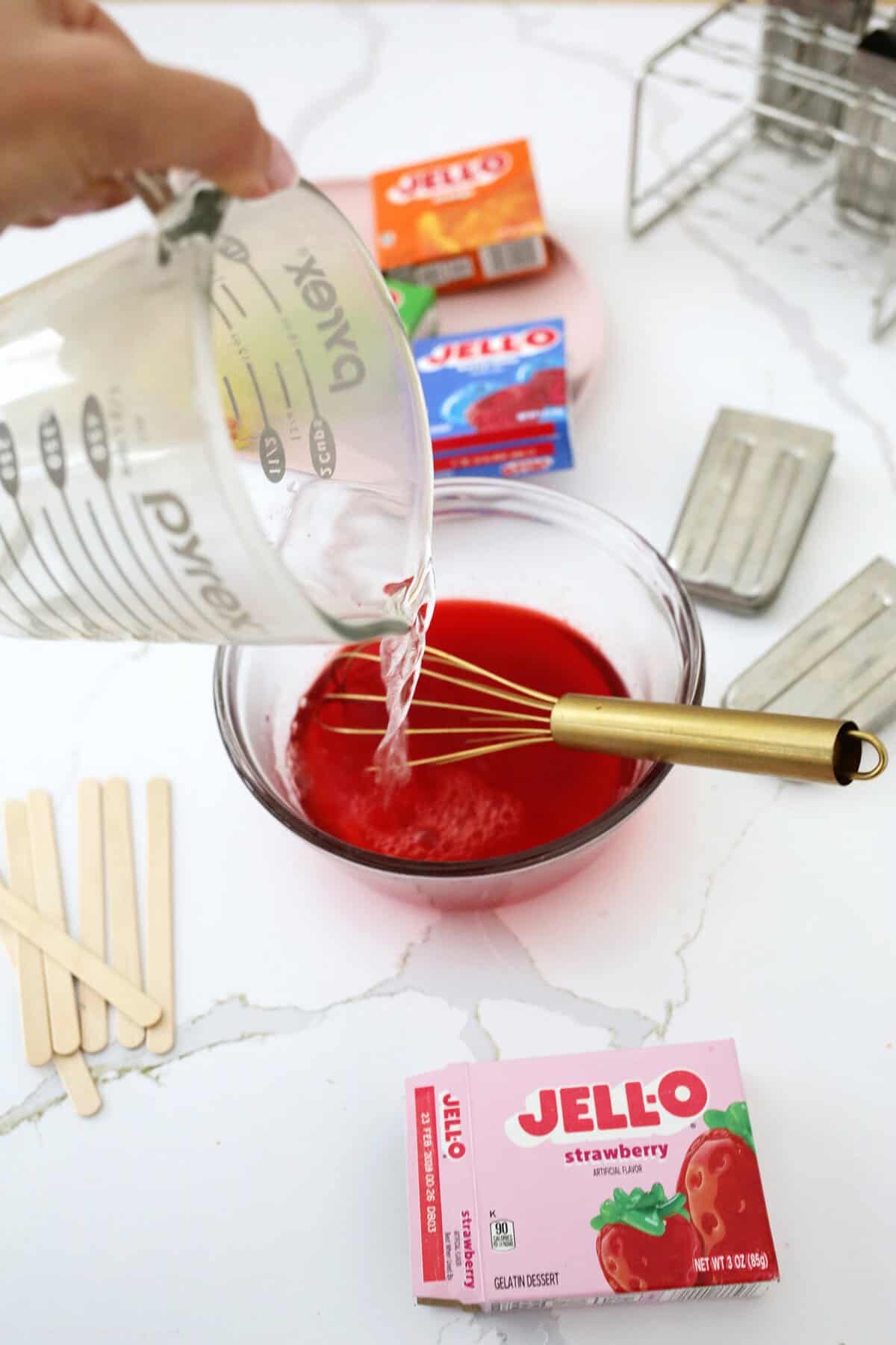 Hot water being added to a clear bowl of red jello powder on a counter next to boxes of jello.