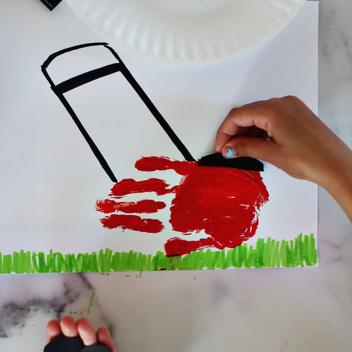 Child adding black cut out paper to a red handprint to make it look like a mower.