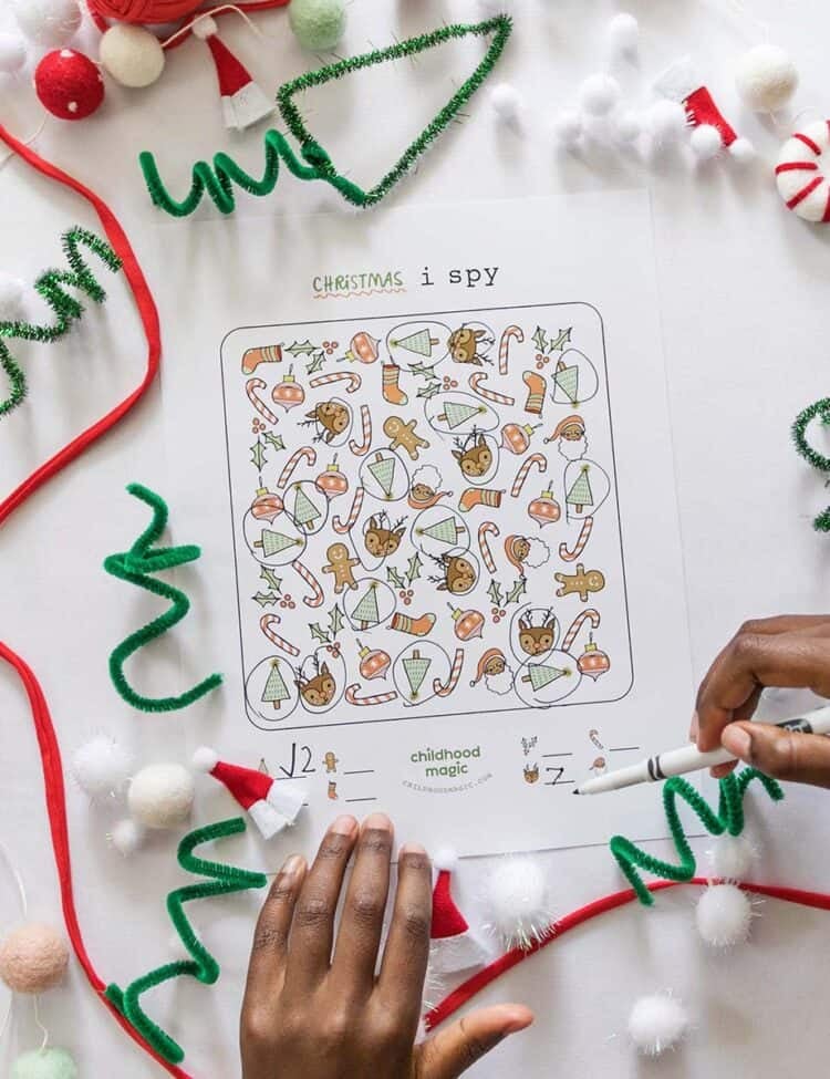 A child's hands next to a Christmas themed I Spy printed game on a counter with pipe cleaners and other Christmas craft supplies.