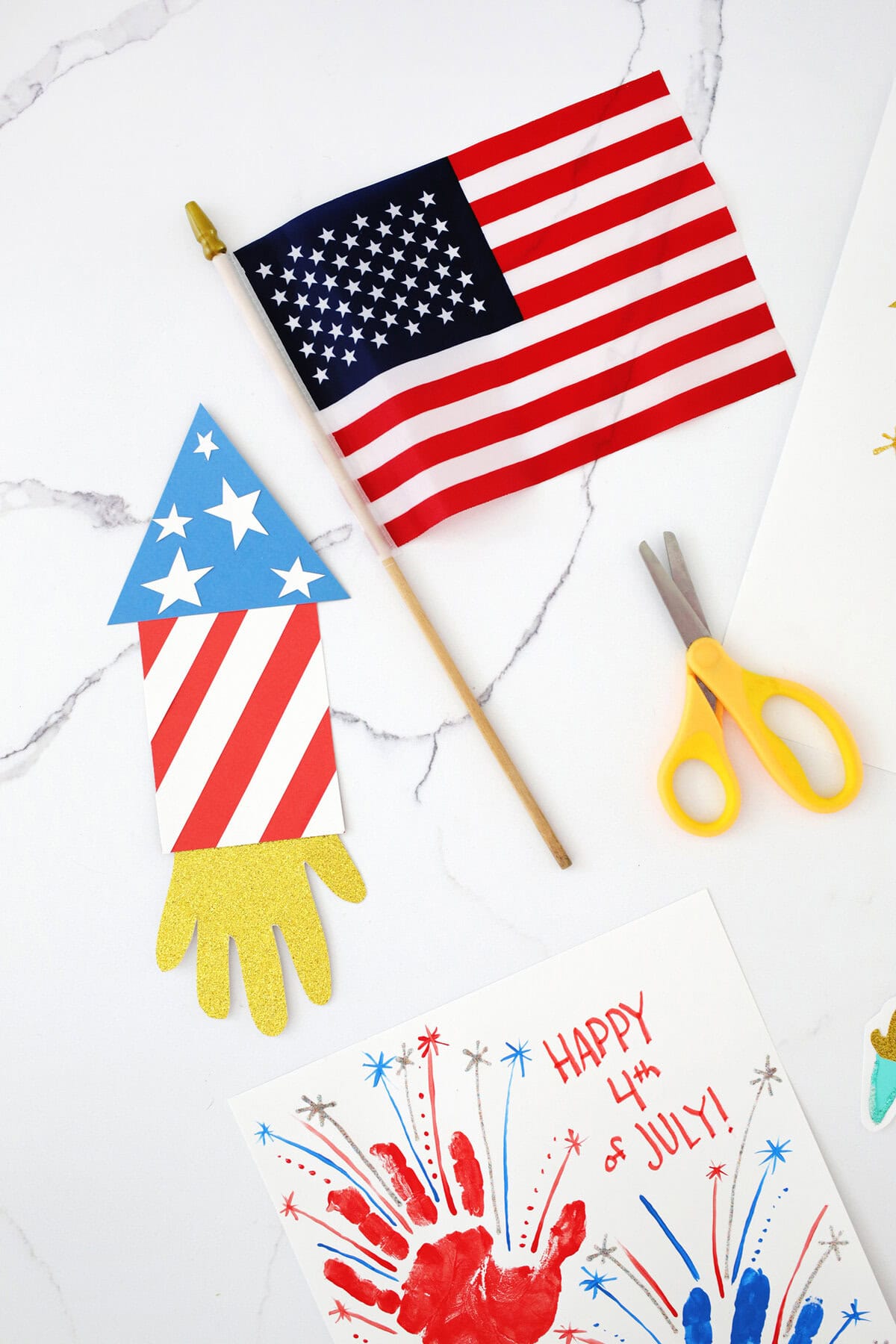 Fourth of July handprint crafts on a counter next to a pair of yellow scissors and a small American flag.