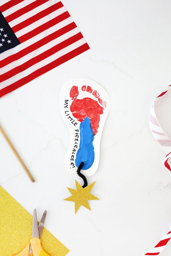 Painted footprint on a piece of paper in red and blue paint with a starburst cut from gold cardstock attached to a piece of yarn and to the footprint.