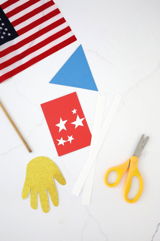 Handprint cut out of gold cardstock next to red, white and blue cardstock cut out in a triangle and rectangle.
