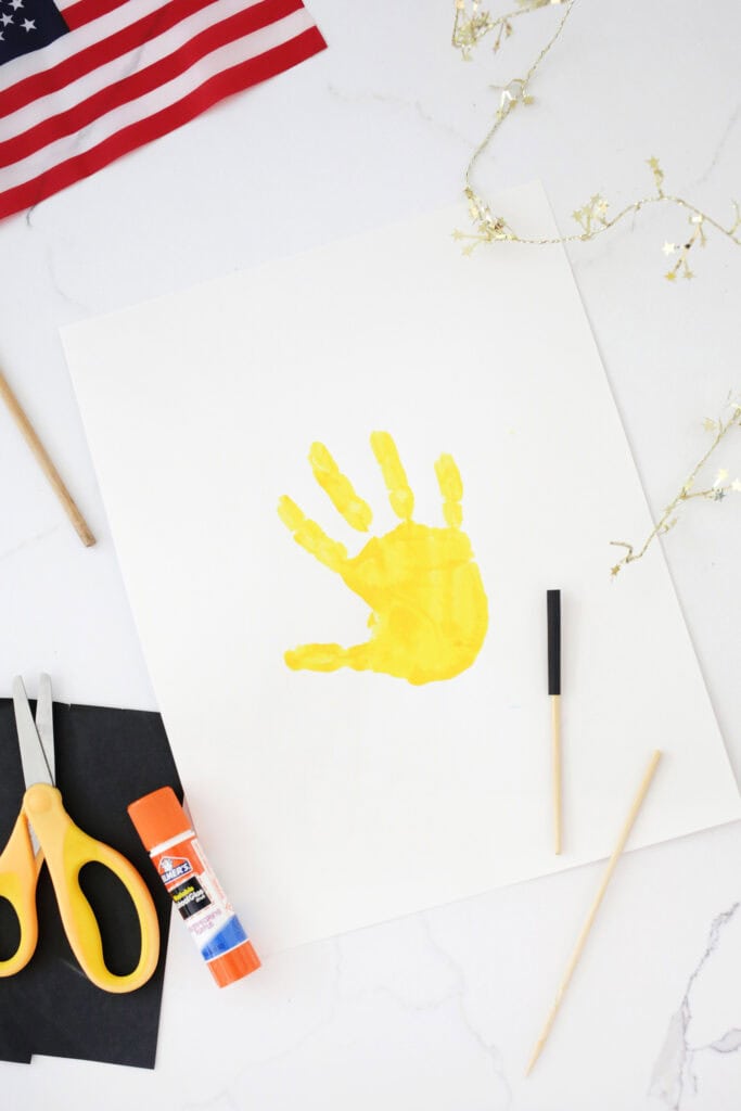 Yellow handprint on a white piece of cardstock on a counter next to a painted stick, glue stsick and pair of yellow scissors.