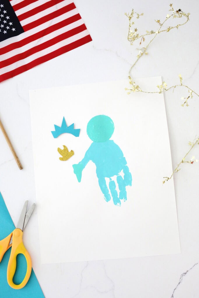 Turquoise handprint on a white piece of paper on a counter with a circle painted near the palm of the hand.Cut out crown and flame next to the handprint.
