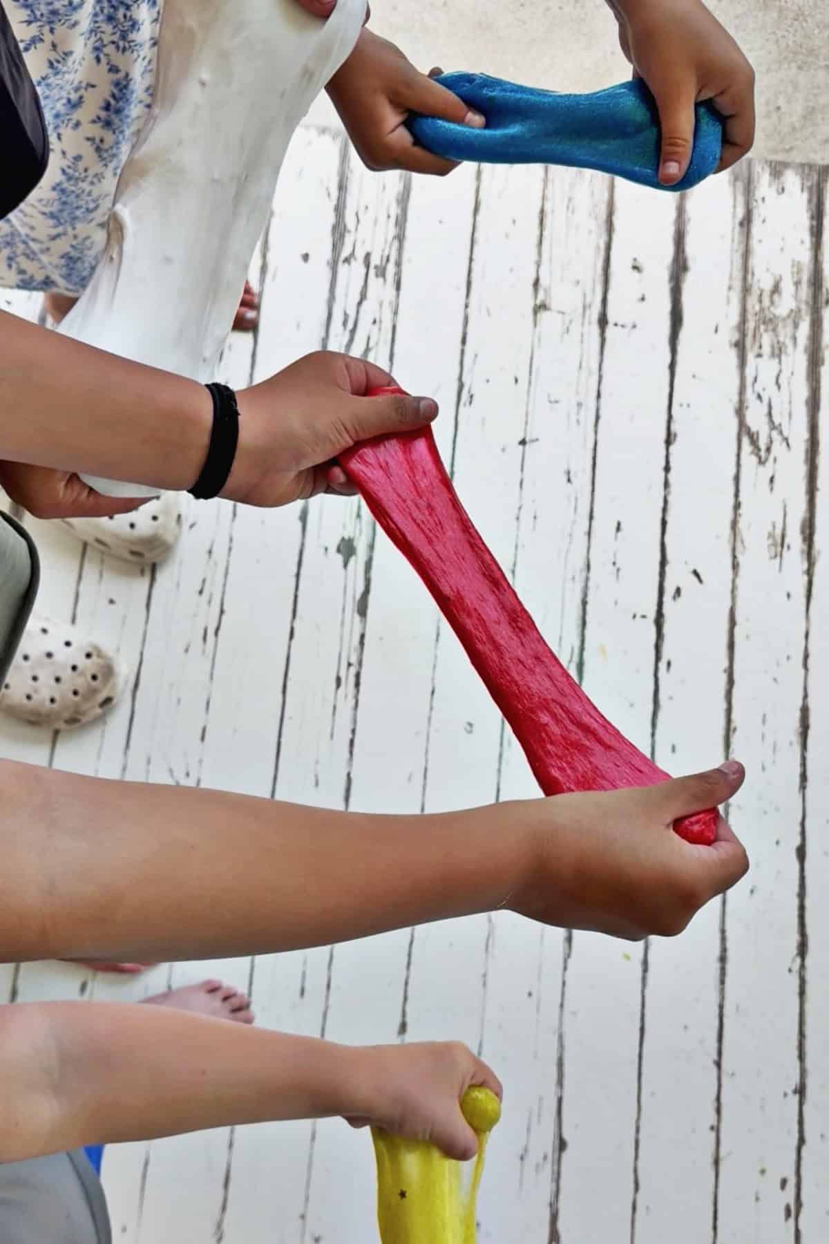 kids hands pulling apart yellow glitter slime, blue glitter slime, red glitter slime and white fluffy slime.