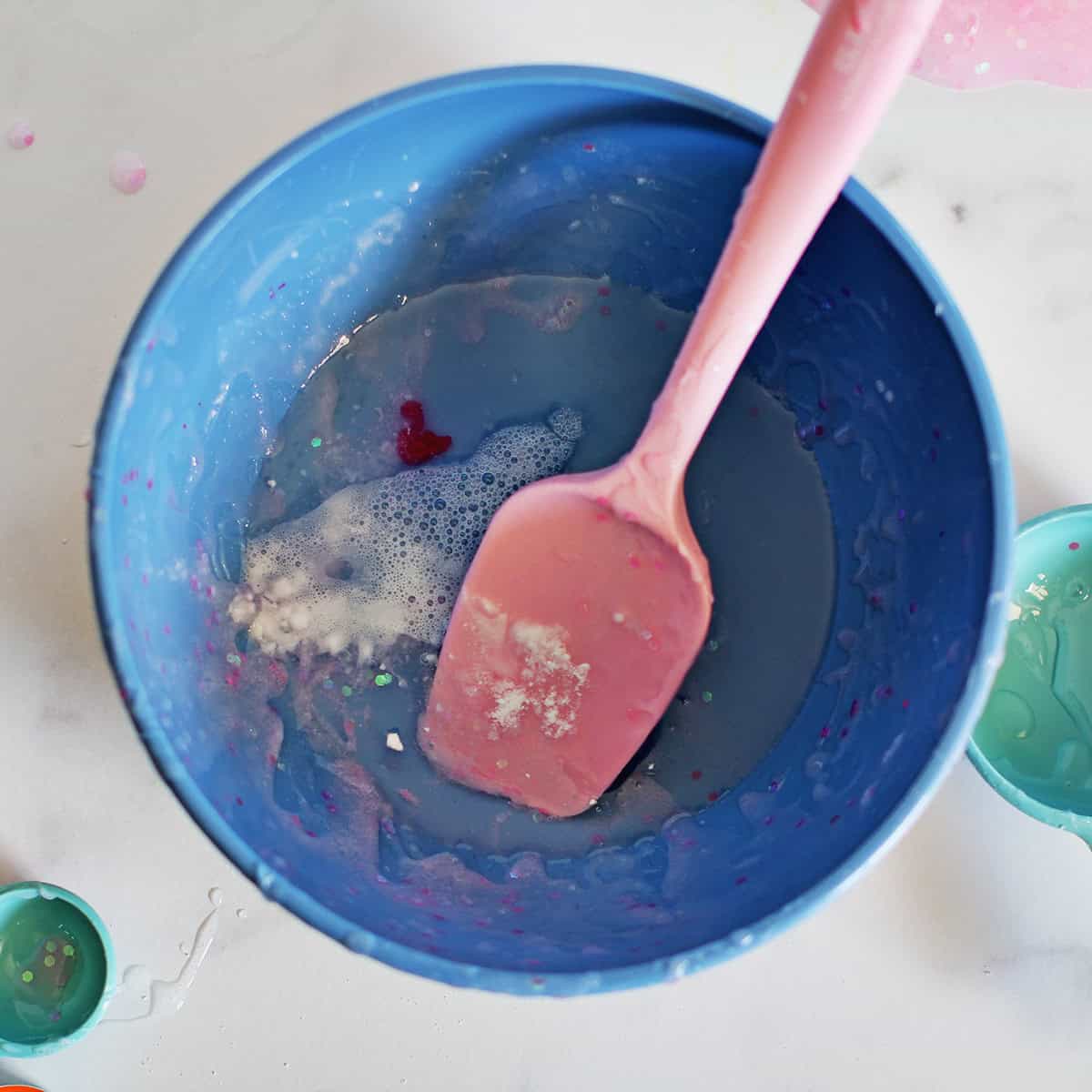 Glitter, contact solution, baking soda and clear glue in a blue plastic bowl being combined by a pink spatula.
