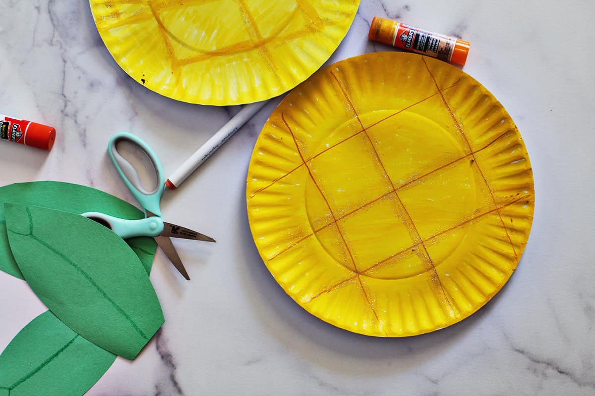Paper plate painted yellow with lines drawn on it to make it look like a pineapple next to green construction paper leaves.