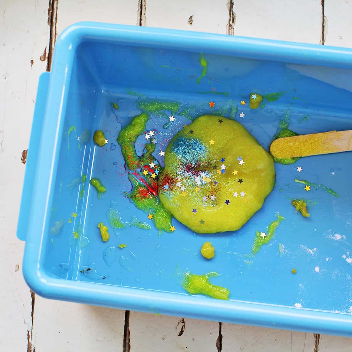 yellow glitter glue in a tub with red and blue glitter, a popsicle stick to stir and gold glitter stars.