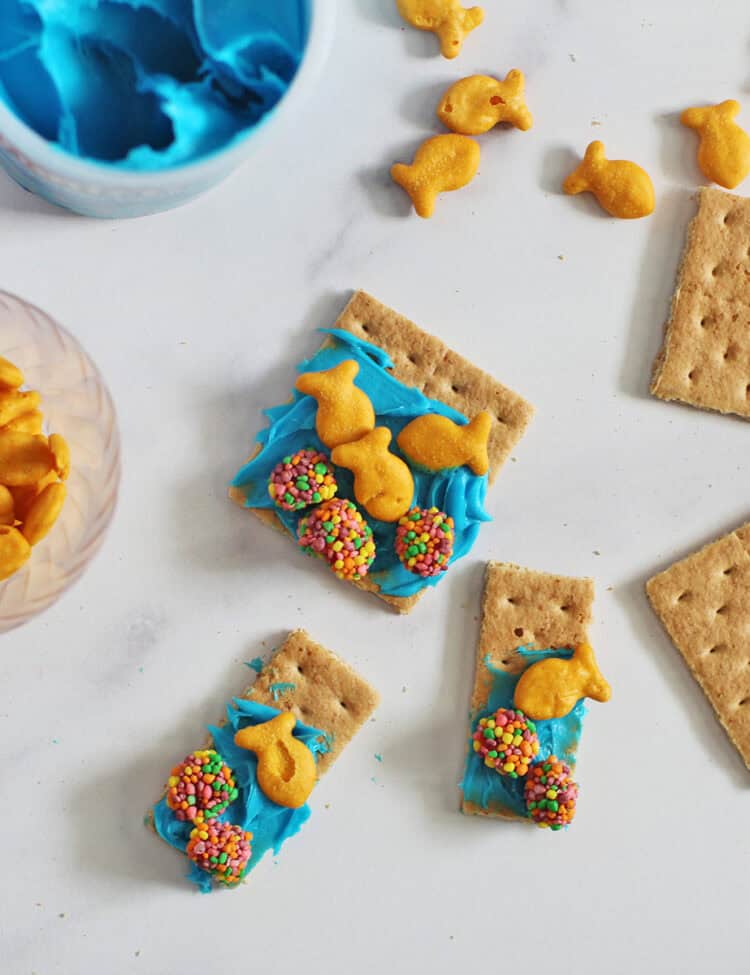 graham crackers with blue frosting, gold fish crackers, and nerd clusters next to a container of blue frosting.
