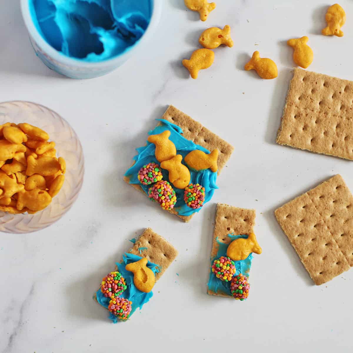 graham crackers with blue frosting, gold fish crackers, and nerd clusters next to a container of blue frosting.