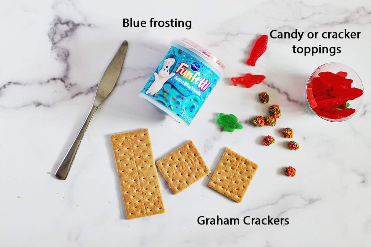 Container of blue frosting, graham crackers, candies and a knife on a counter.