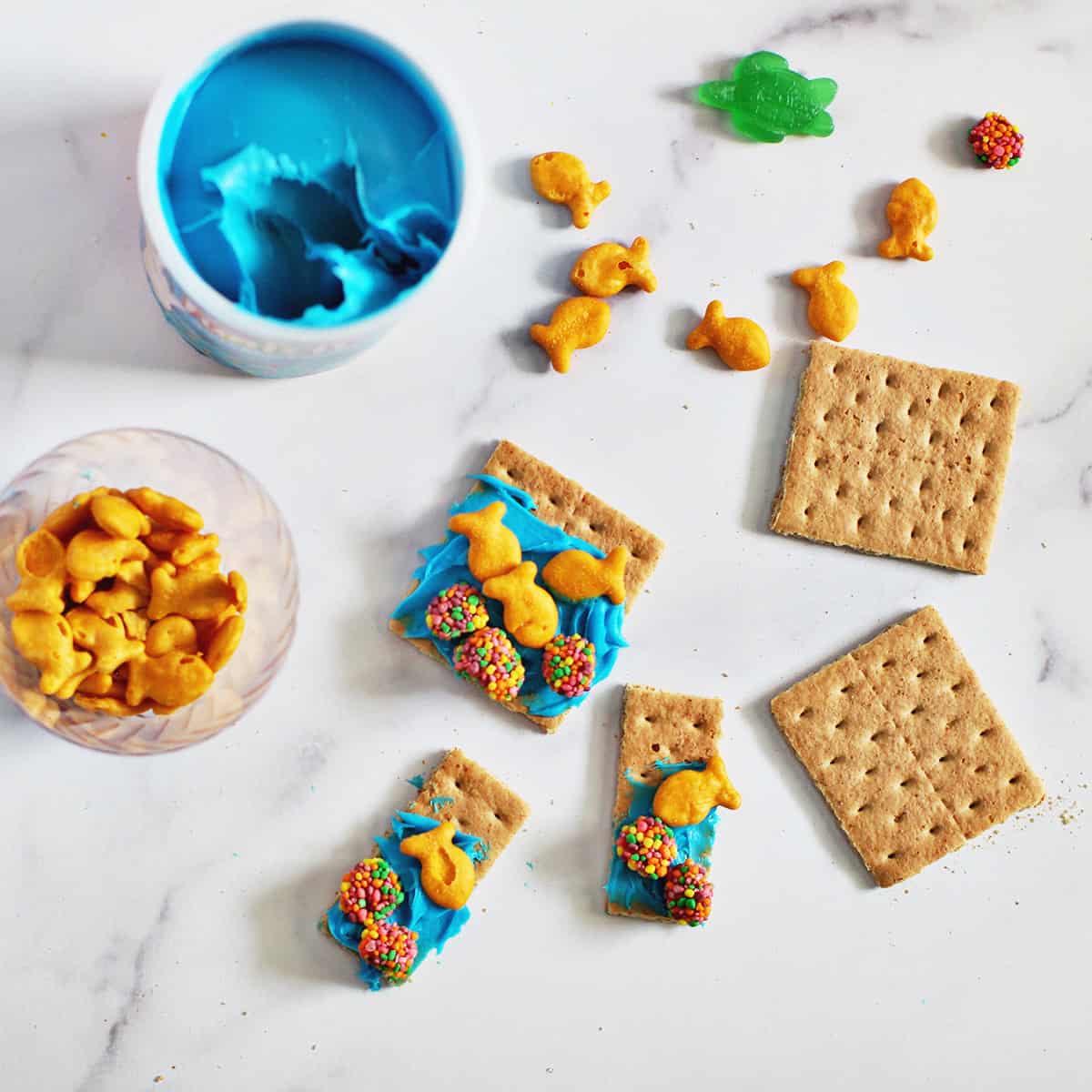graham crackers with blue frosting, gold fish crackers, and nerd clusters next to a container of blue frosting.