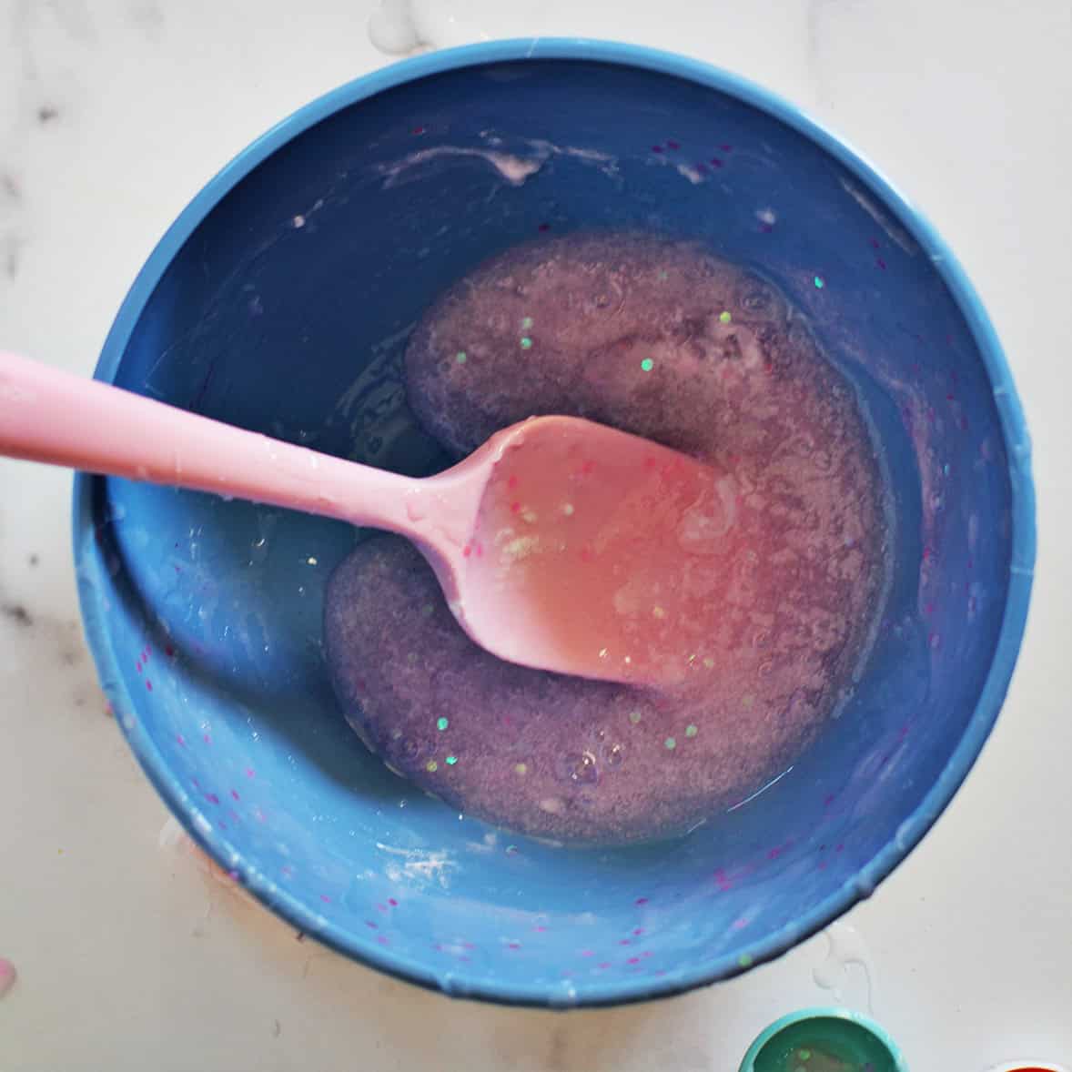 Pink glitter slime being combined in a blue bowl with a pink spatula.