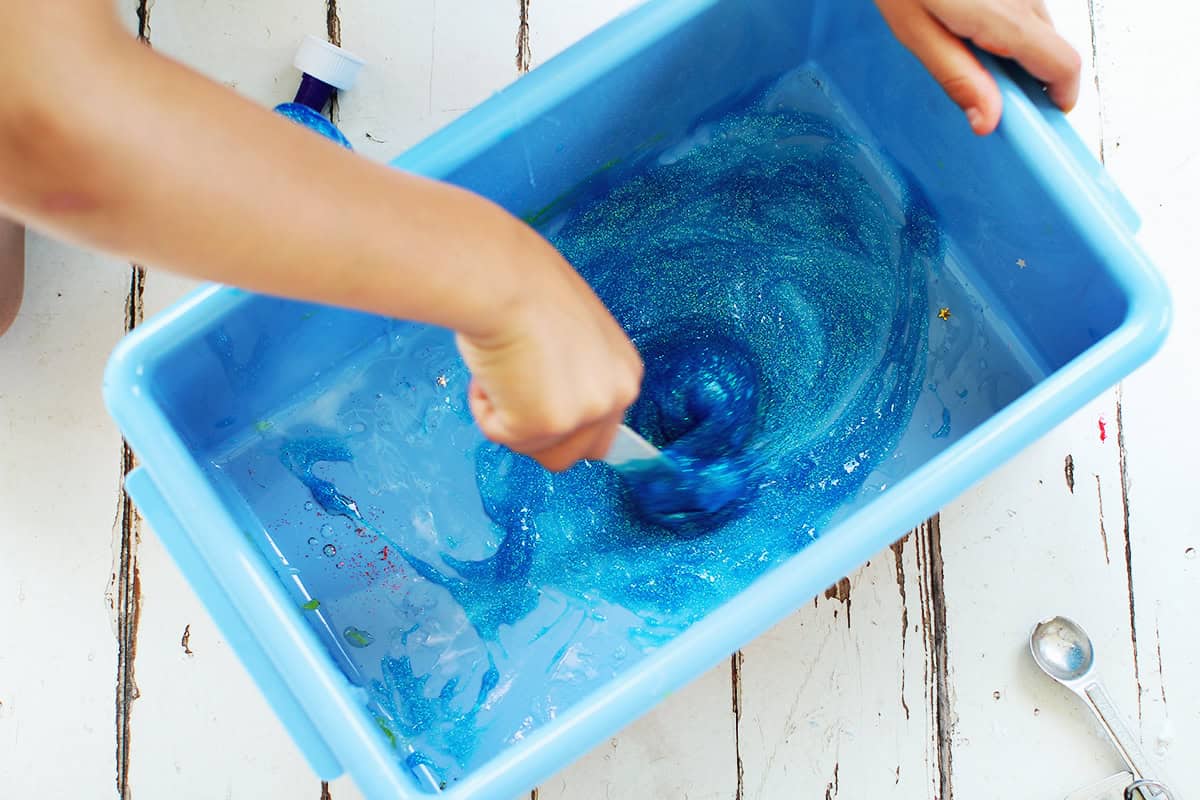 Child's hand stirring blue glitter glue with contact solution to make slime.