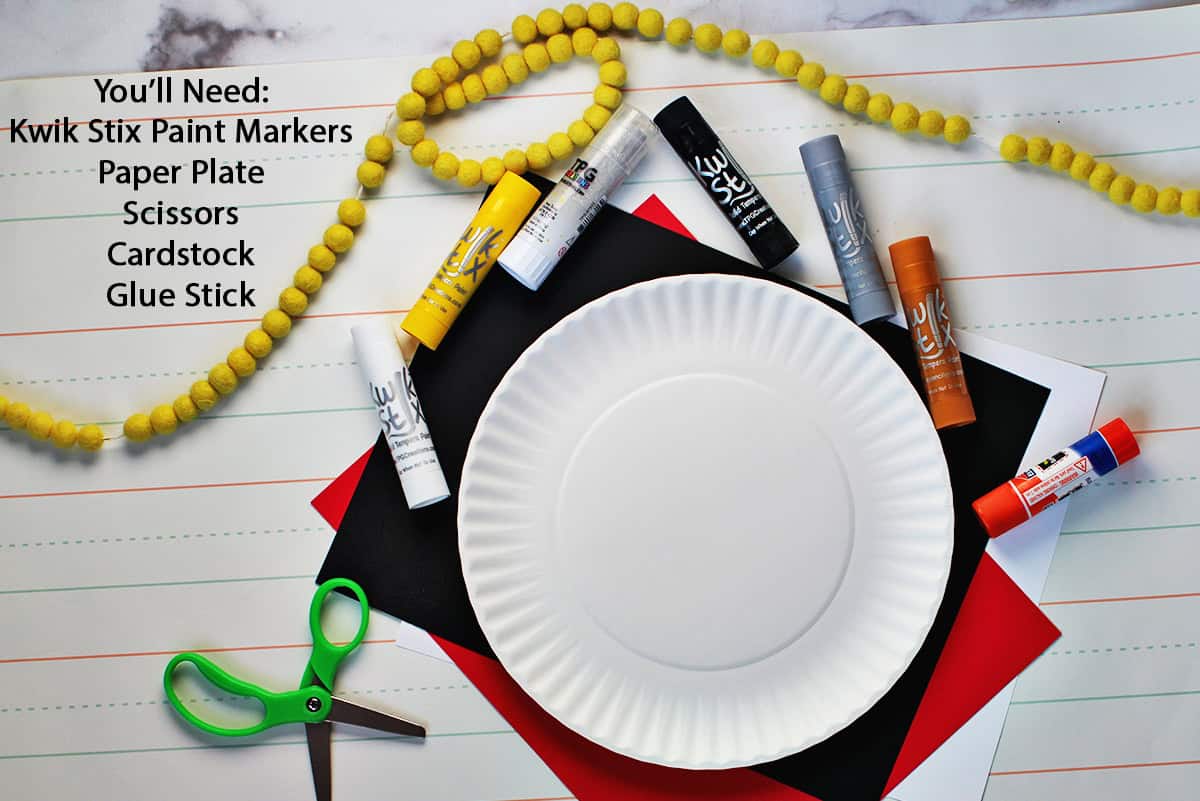 Plain paper plate, green scissors, a glue stick and paint pens on a counter.