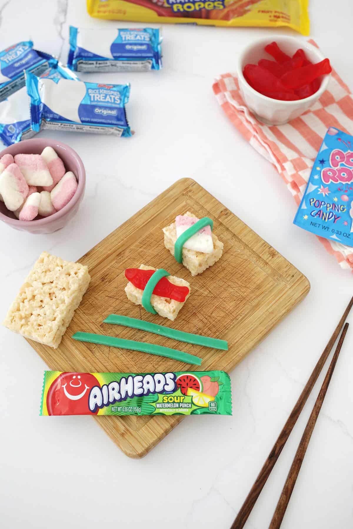 sushi candy being made on a cutting board with swedish fish, a rice krispie treat and a green airhead.