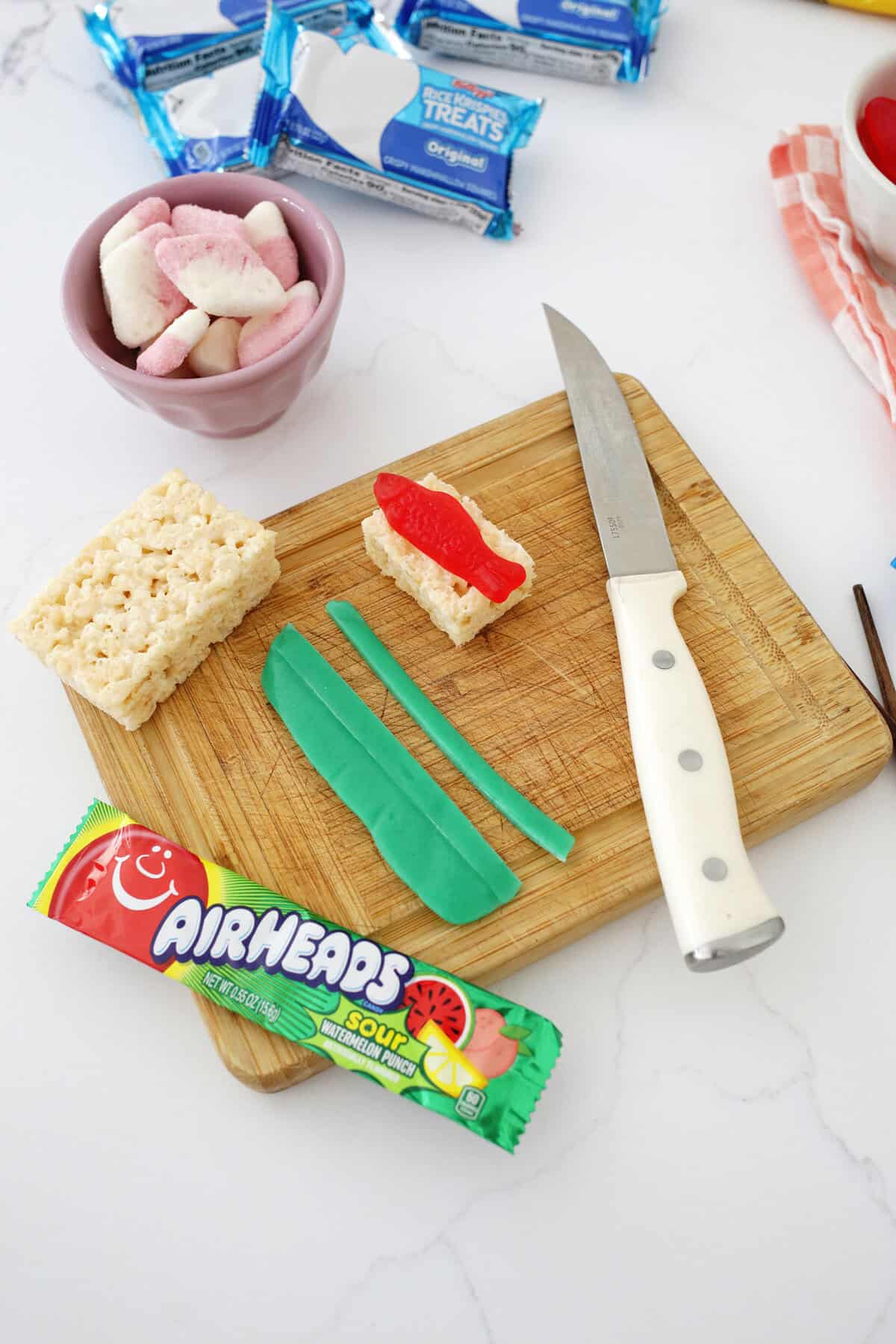 A knife on a cutting board next to a green air head, rice krispie treats and a swedish fish making a candy sushi roll.