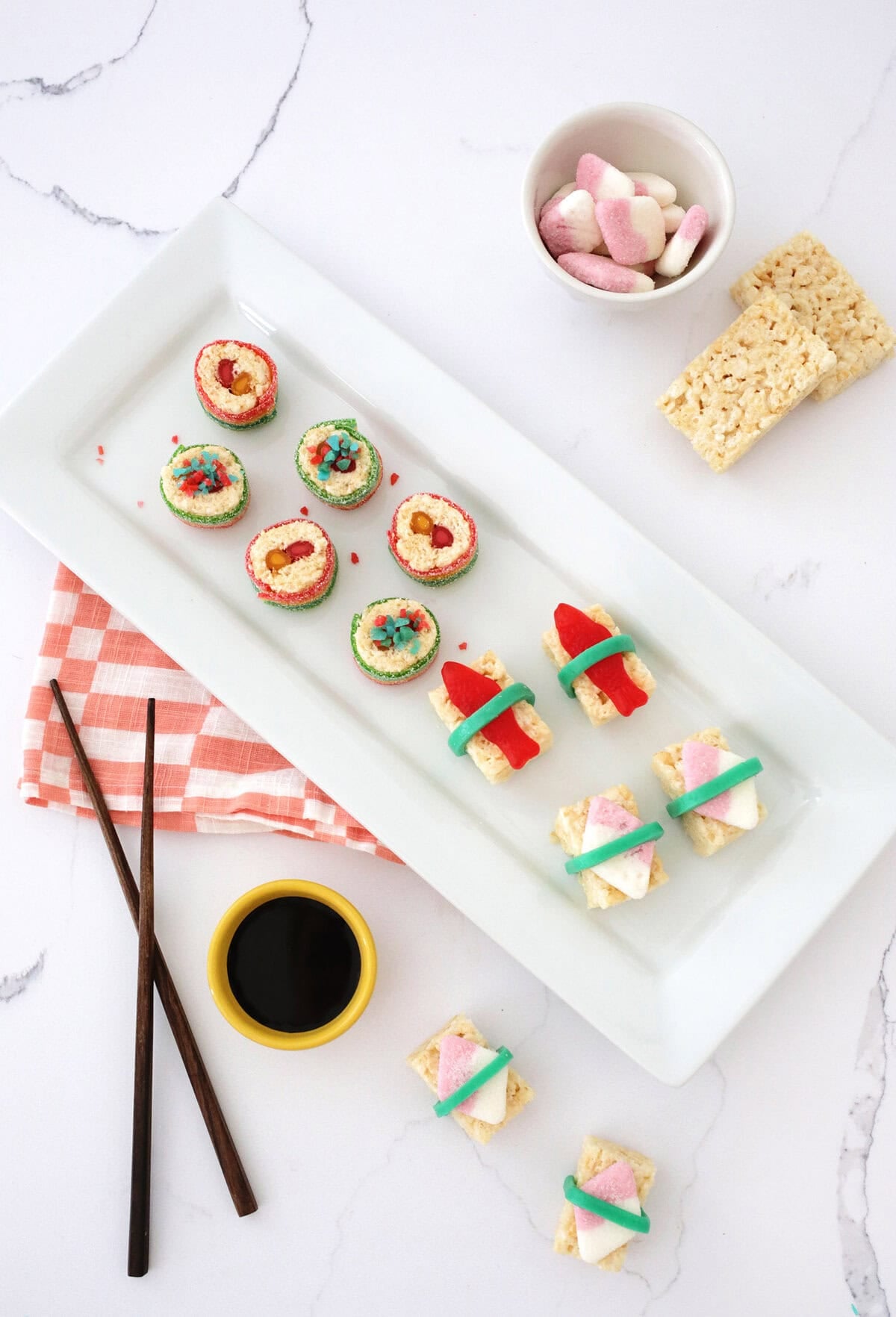 candies made to look like sushi pieces sitting on a white plate next to chop sticks and a small bowl of chocolate syrup.