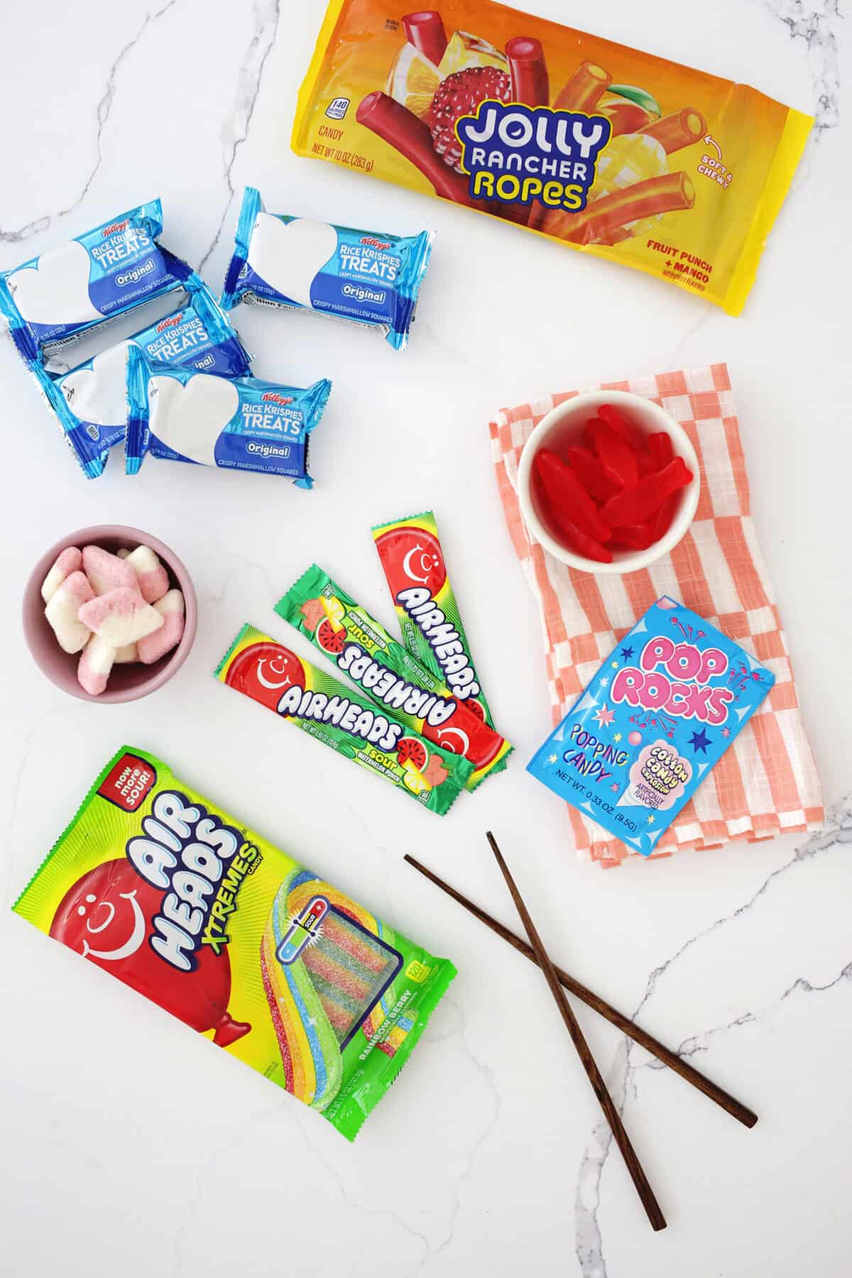 Rice krispie traets, jolly rancher ropes, swedish fish, airheads and other candies on a counter.