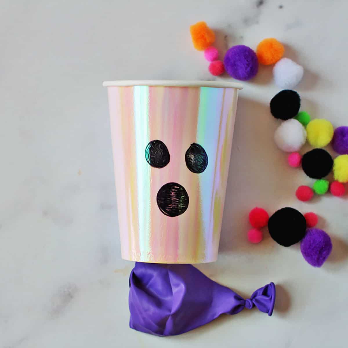 cup with a ghost face drawn on it next to a cut balloon and pom poms.