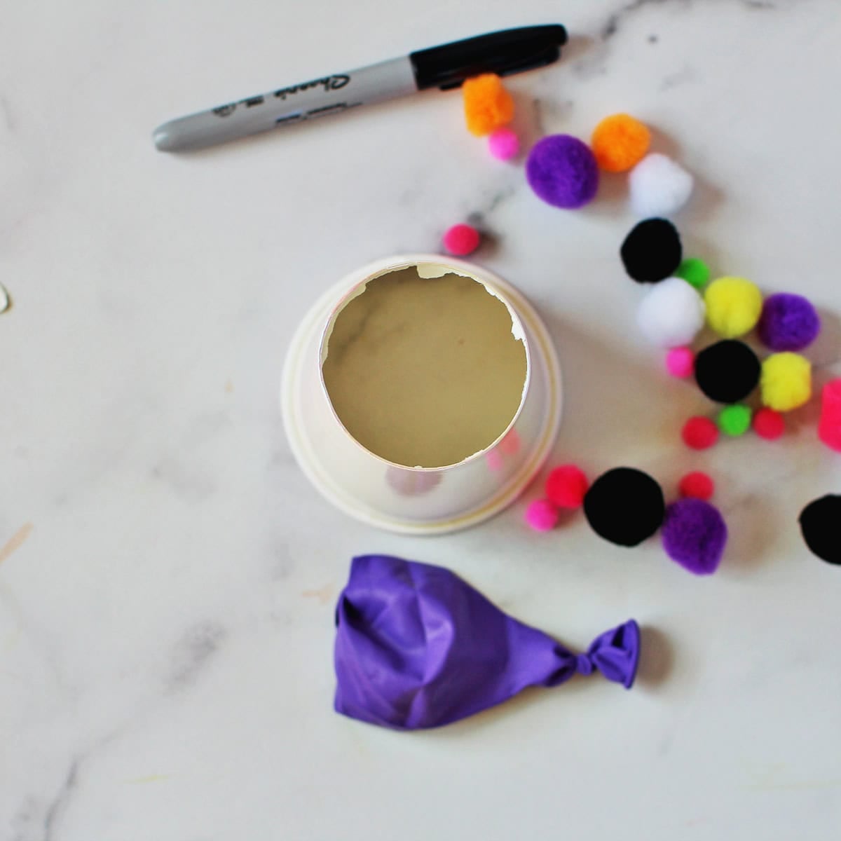 White cup with bottom cut out of it next to a purple balloon and pom poms on a counter.