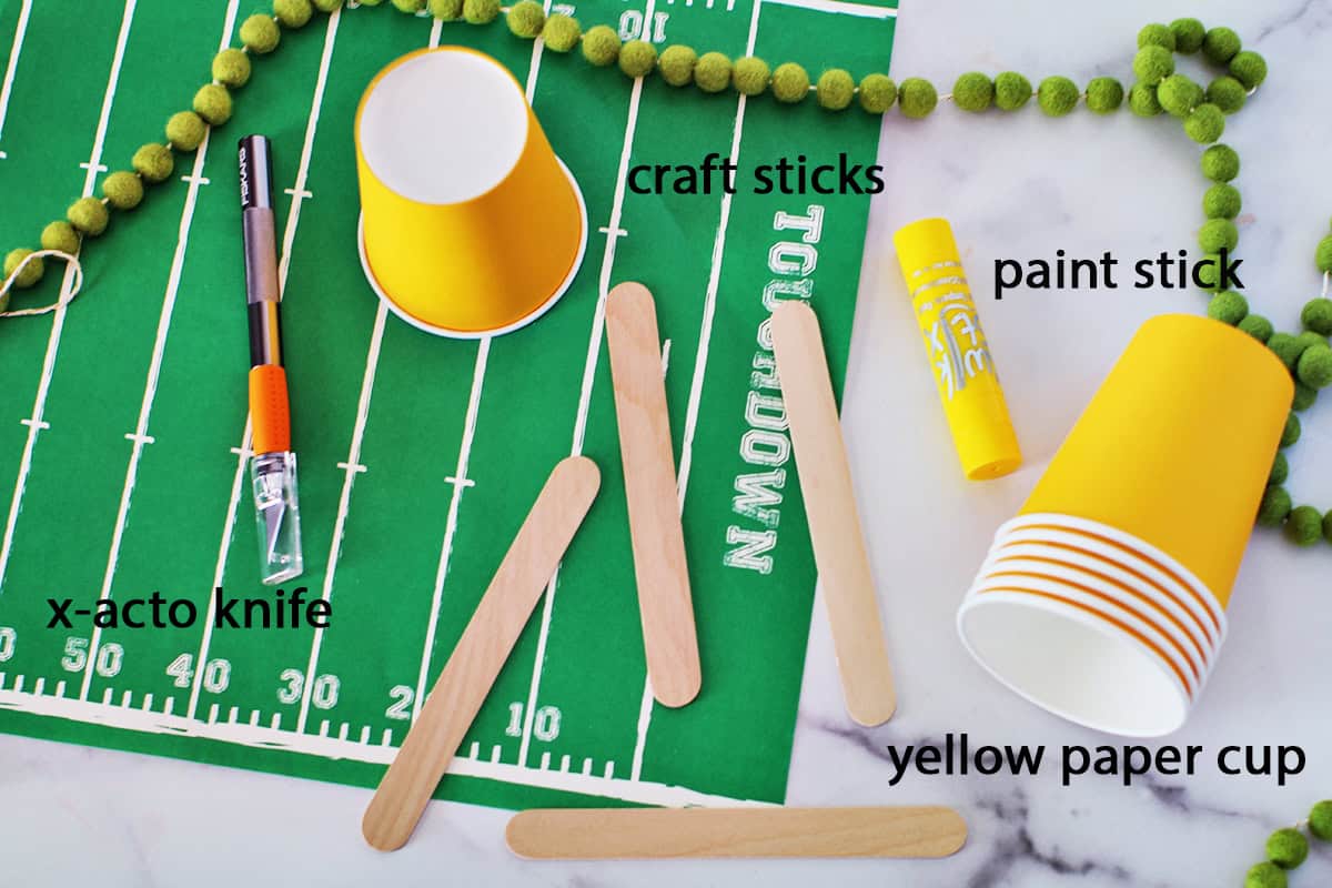 Yellow paper cup, craft sticks, an x-acto knife and a paint marker sitting on a placemat that looks like a football field.
