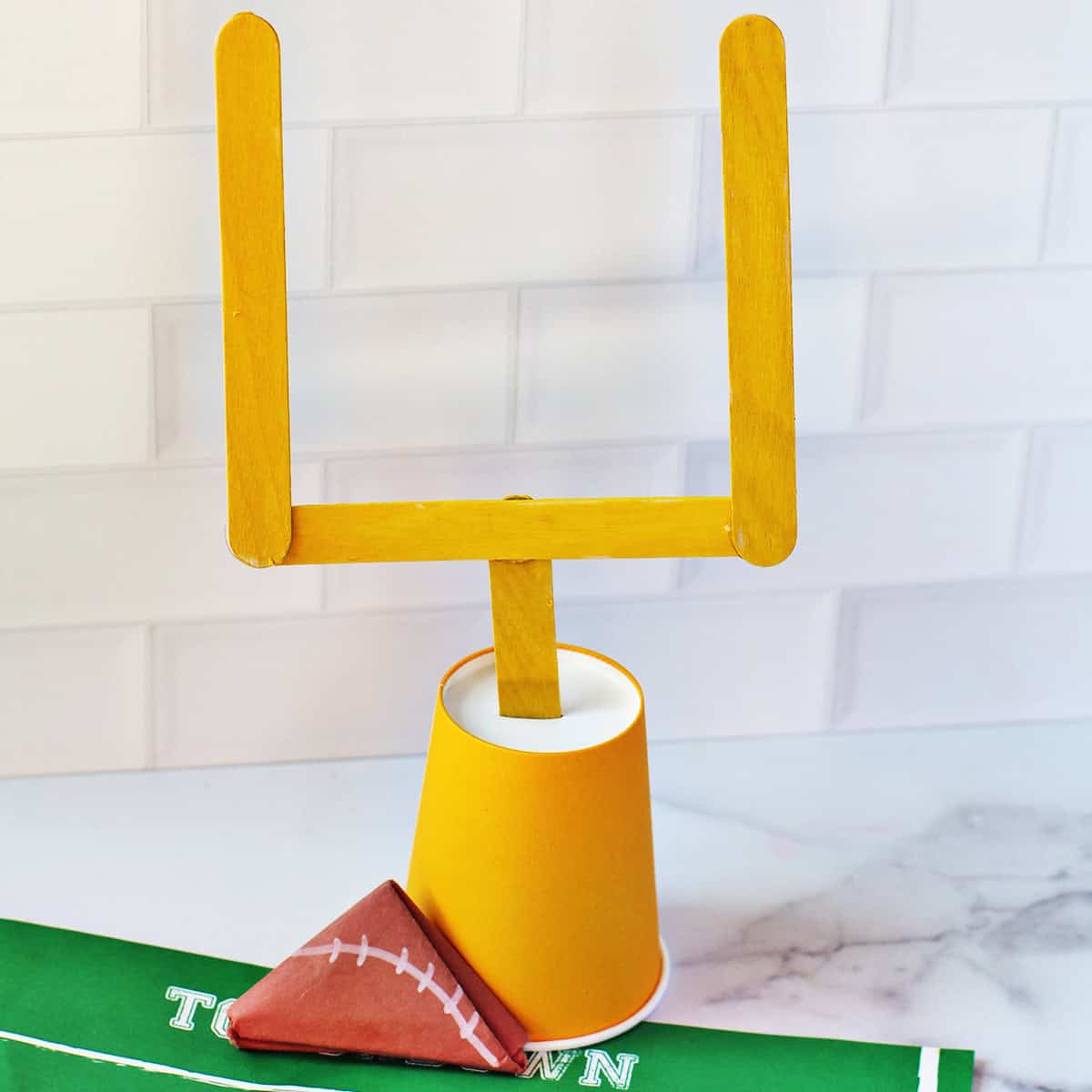 Football goal posts made from yellow craft sticks and a cup next to a paper folded to look like a football.